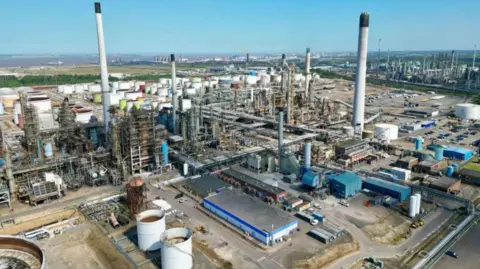 BBC A drone view of the Prax Lindsey Oil Refinery. A cluster of metal structures rising to the blue sky.