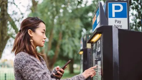 A woman with long brown hair and a jacket uses a parking payment machine and has her phone in her left hand. She is tapping buttons on the machine and looking thoughtful. There is a park behind her.