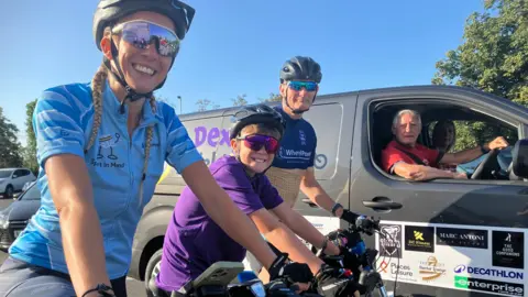 From left to right - Dexter's mother Sam, Dexter and coach Dave Covey as smiling as they are prepared to set off. All three are on their bikes. Sam wears a blue top for Sport in Mind, Dexter wears a purple T-shirt for Falconi Hope, and Mr Covey wears a dark blue and yellow top for WheelPower. A driver in a van with Dexter's name on it is next to them. A woman can be seen in the passenger seat. It's a sunny day.