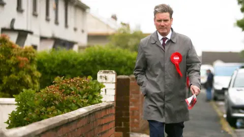 Keir Starmer walking on a residential street towards the camera in 2017. He is wearing a red labour rosette and a rain mac