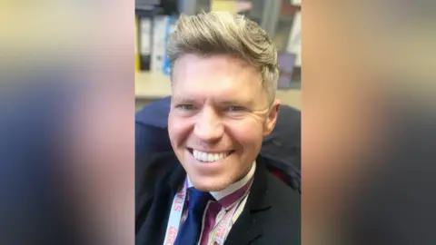 Brian Murphy Brian Murphy is sitting in an office. He has blond hair that's styled in the front. He's wearing a black jacket and a navy tie. A lanyard saying staff is around his neck. 