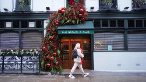 A woman is seen walking past the Groucho Club entrance. The wooden doors with windows have green awning with the club's name and it is decorated with a Christmas tree beside it