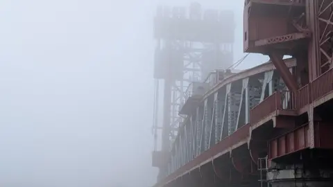 TheAdder/ Weather Watchers Newport Bridge in Middlesbrough. It is a brownish red steel structure with grey beams. Fog surrounds the higher parts of the bridge and creates an entirely grey background against which the colour of the structure stands out.