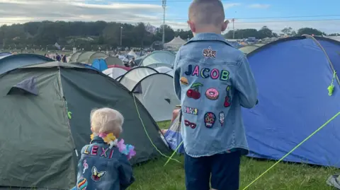 Family handout Two children with their backs to the camera stood amongst a number of tents. They are wearing their festival denim jackets with their names on and decorated with pictures including donuts, butterflies, cherries, flamingo's, and a skull.