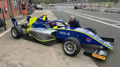 The Idola Motorsport GB4 car which is blue with a bright green and silver patten. Jamie is sitting in the car. 