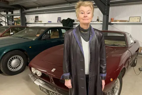 A woman with short blonde hair, wearing a long dark leather coat, stands in front of a sporty-looking maroon car.