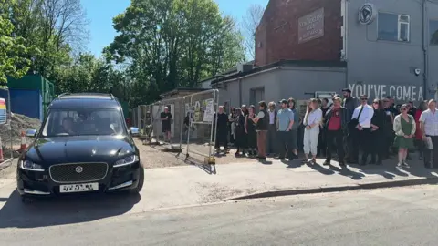 A black hearse pulls out if the car park of the New Adelphi Club a crowd stands outside the venue watching