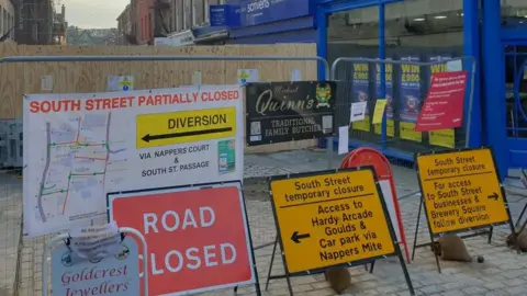 Multiple signs in front of a wooden fence that's sealing off part of the shopping street. One has a map and reads South Street partially closed, diversion. Another says road closed. Others mention specific businesses and have arrows to direct people for access.