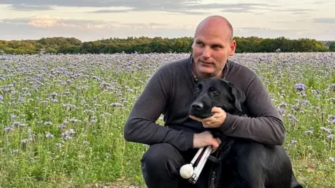 BBC correspondent Sean Dilley pictured in a field on a bright day, crouched down with his arms around his black guide dog Shawn