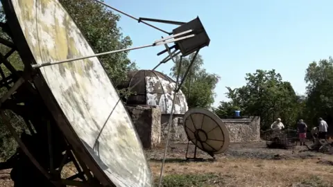 A large telescopic dish stands abandoned and dirty at the site with other old equipment dotted about while people work in the background.