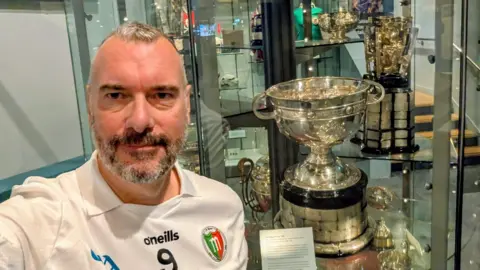 Sean McAndrew A man wearing a sports jersey standing in front of a glass cabinet with a large silver cup inside it.