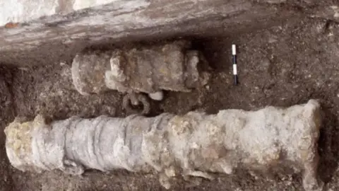 Humber Field Archaeology An archaeological photograph of a excavated cannon lying in a ditch which is in two parts and is covered in rust and earth