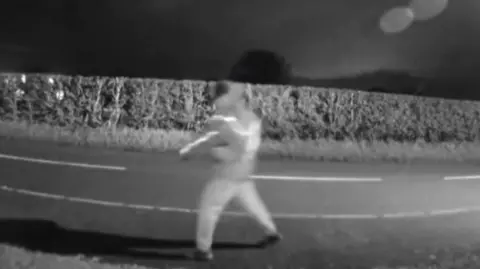 A blurred image of a man walking past a house at night with the road in the background. The image was taken from Ring doorbell footage.