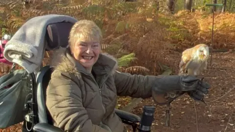 Kim Smith A woman in a dark green coat, holds an owl with her gloved left hand.
