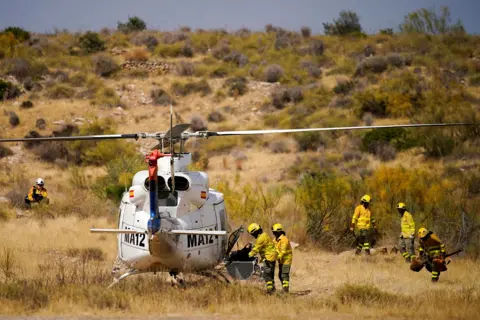 Ana Beltran / Reuters Firefighters in yellow hi vis and helmets gather around a helicopter parked on shrubland in Almocaizar on 12 July.