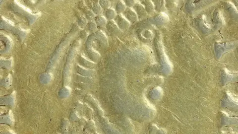 Andrew Williams/Norfolk County Council A close-up of an Anglo-Saxon gold coin. It shows an attempt to replicate the face of a Roman emperor in profile and is quite crudely made