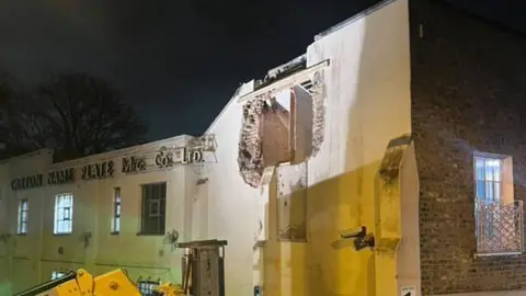 A night time scene of a partial building with a large hole exposing bricks and wooden framing. The faded signage reads "Mfg. Co. LTD." A boom lift is visible below, and artificial lighting casts shadows against the night sky.