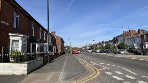 A road with red brick terrace houses with small front gardens and cars parked along the kerb.