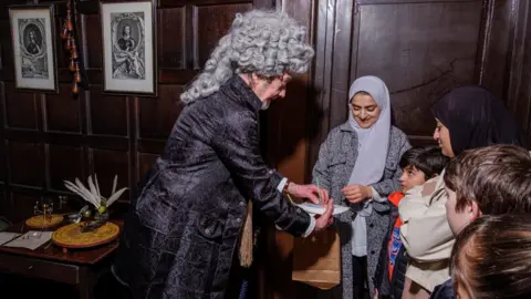 Bradford Museums/Phil Jackson A man in a grey curly wig and velvet coat speaks to some children and shows them some writing