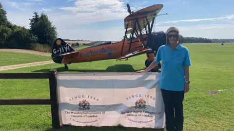 Catherine Grimley Catherine Grimley looking at the camera with the RFC Rendcomb field and a biplane behind her. Mrs Grimley is wearing a brown hat, blue t shirt and black trousers.