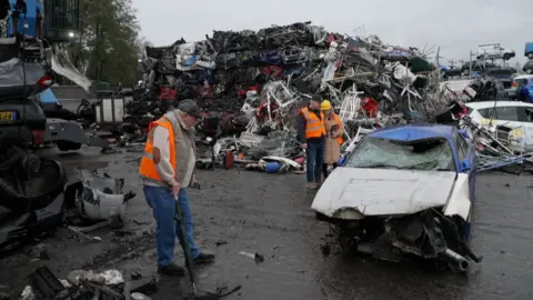 Three people in a scrapyard looking at a smashed-up car