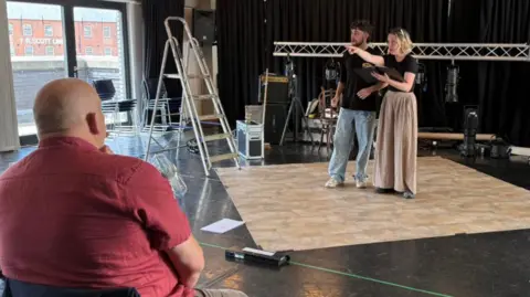 The photo shows John sitting to the left of the photo. He's wearing a red shirt, and is facing away from the camera. He's looking at two people who are standing on a stage. They're acting out a scene from a play. There are various stage props, such as lights and ladders, also scattered around the room. 