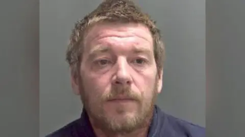 Custody photo of Wayne Freeman against a grey background. He has ginger hair and a ginger beard. Freeman wears a navy blue sweatshirt and is looking to the side of the camera and not smiling. 