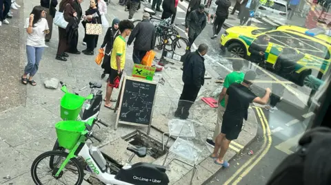 Paul Barlow / X Image taken from what appears to be upper deck of a bus shows people gathered on a street corner, some watching while others walk by, there are Lime rental bikes in the foreground and an emergency vehicle in the road in the background