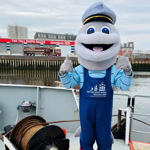 Dorry the dolphin mascot on a boat, giving thumb up, with sign saying 'Tall Ships Races Aberdeen 2025' in the background.