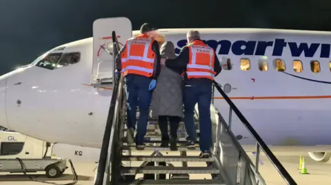 Two men in orange high-viz vests with back-up team written on the back lead a woman in a grey coat and dark trousers up the steps of a plane