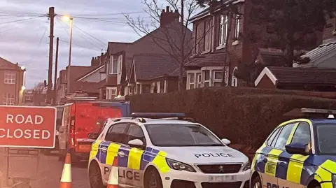 BBC Two police vehicles parked in a cordon on Salisbury Avenue, Blackpool, next to a red and white 'road closed' sign.