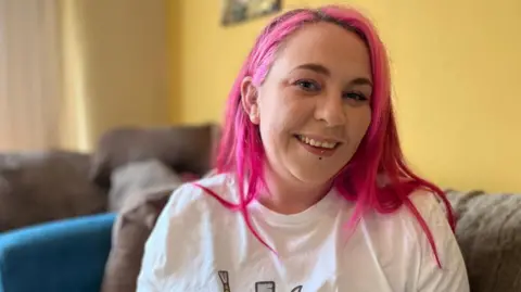 Jenny has long, bright pink hair and is wearing a white t-shirt. She is smiling at the camera and is sat in a yellow living room on a blue and brown sofa.