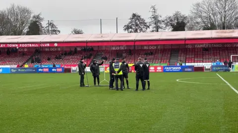 James Dunn Six people in all black clothing stood in the middle of a football pitch. In the background are red seating stands