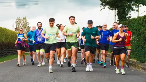 Laura-Mary Carter/FTD Brothers Runners going from left to right on the photo are all facing the camera. Bushes are either side of the road that they are on.