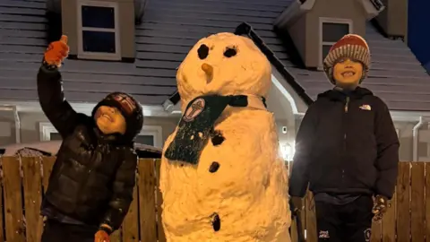 Two boys in hats, jackets and gloves beside a snowman outside a house in Feeny