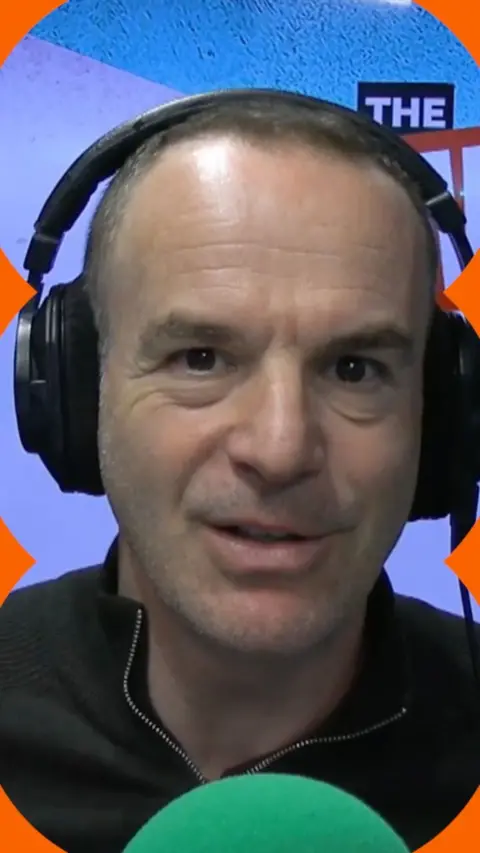 Martin Lewis wearing black headphones and zip top, sitting behind a green mic