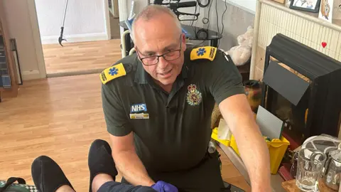 A man in a green ambulance uniform kneeling on the floor checking on an elderly man who is sitting in a chair