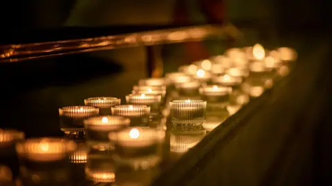 A stock image of lit remembrance candles in glass tealight holders on a shelf in a dark church.