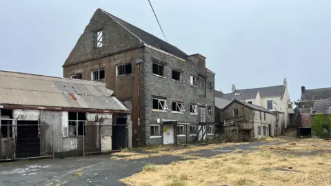 Part of Leale's Yard which has five derelict buildings in a row and another adjacent. the largest one has four floors. Its window frames are open but there is no glass in any of them. There is dead grass in the courtyard-like area in the foreground.