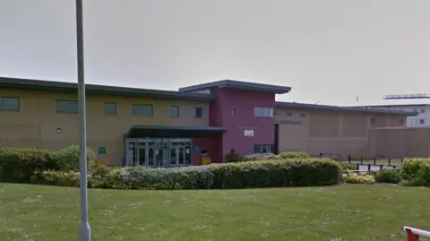 Google Street view image of the prison from behind a barrier. It is a long rectangular building with a red on the walls where the entrance is. Outside is a stretch of grass with some bushes and flowers.
