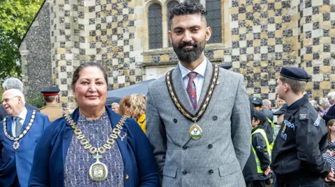 A picture of Naheed Ejaz, wearing her mayoral chains, and her son, Diwan Khan, wearing the consort's chains, outside an event with people milling behind them. 