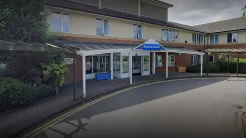 Google Parklands Hospital, with a white awning over its entrance, a blue sign and a road running outside and curving away from it.