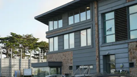 Exterior of La Moye Prison in Jersey. It has grey cladding on the outside of the building. It has several windows and a couple of cars parked outside. Trees are in the background behind a large white fence.