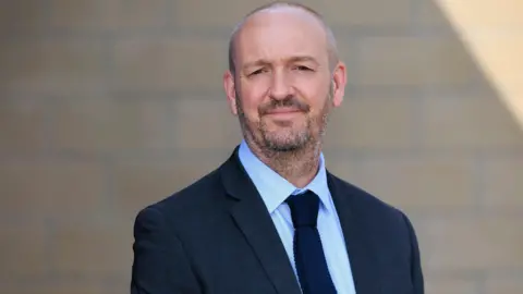 GMP David Sinclair, who is bald has black and grey stubble, looks into the camera wearing a navy blue suit jacket over a light blue shirt and dark blue tie