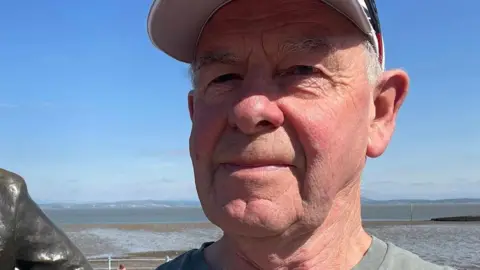 A selfie-style image of Jonathan Huggins. He is wearing a cap and a grey T-shirt. He is standing outside, seemingly on a coastline, while smiling and looking into the camera.