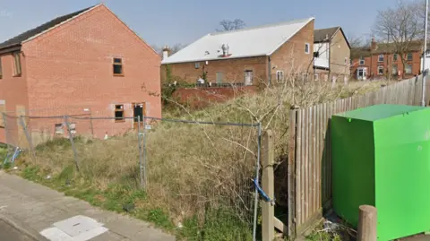 An empty site covered in long grass. It has a wooden fence to the right boundary and a silver wire fence to the left of shot.