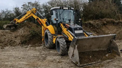 A yellow and black digger parked up by a mound of earth. It has soil in its bucket on the front of the vehicle.