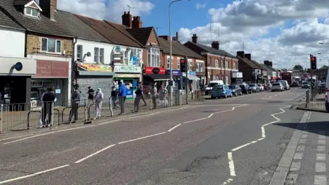 SHARIQUA AHMED/BBC Lincoln Road stretches ahead with white road markings. On the far side is a line of shops and other businesses, with several people leaning against a silver metal gate in front of them. In the distance, cars are parked along the road. A pedestrian crossing bisects the road, with traffic lights showing red in front of it.