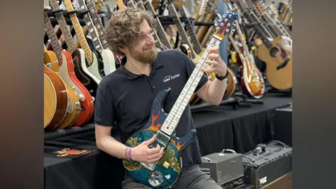 PA A man holding an electric guitar, with his fingers placed on the strings. The guitar is decorated with what looks like a sea with islands and the snow globe is just about visible at the bottom. There are rows of dozens of guitars behind the man