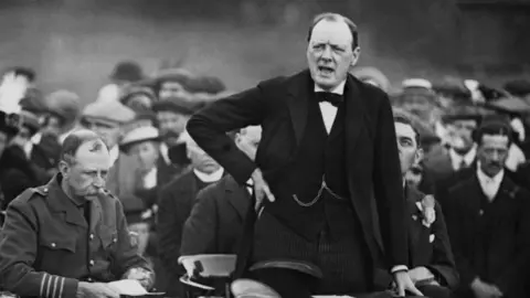 Sir Winston Churchill, wearing a tuxedo, addresses a crowd from a raised stage. He has one hand on his hip.
